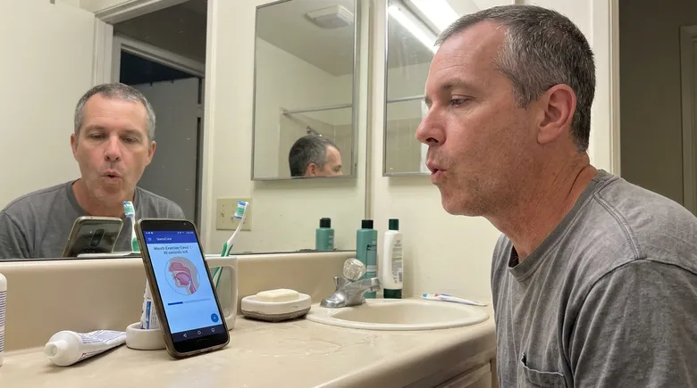 SCENE: Man in his early 50s at bathroom sink, phone propped on the counter showing a SnoreCare exercise screen, performing a morning throat exercise with calm, focused expression