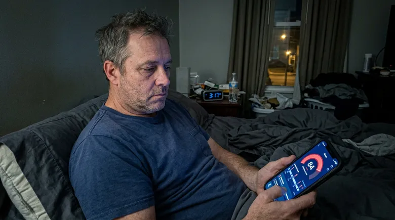 SCENE: A man in his late 40s sitting up alone in bed at 3:17AM, face lit by a glowing phone screen showing a red snoring app score of 84, expression hollow and resigned