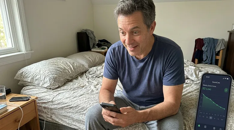 SCENE: Man in his early 50s sitting on the edge of his bed in morning light, holding smartphone showing a steadily declining snore score graph, his expression shifting from skepticism to quiet, surprised relief